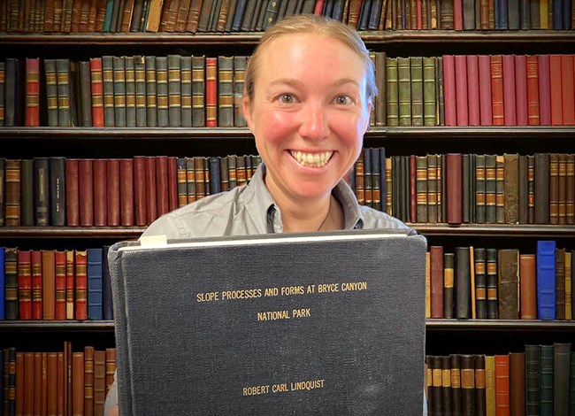 A woman holds a large, gray, old-looking hardcover book in front of a wall of other old books.