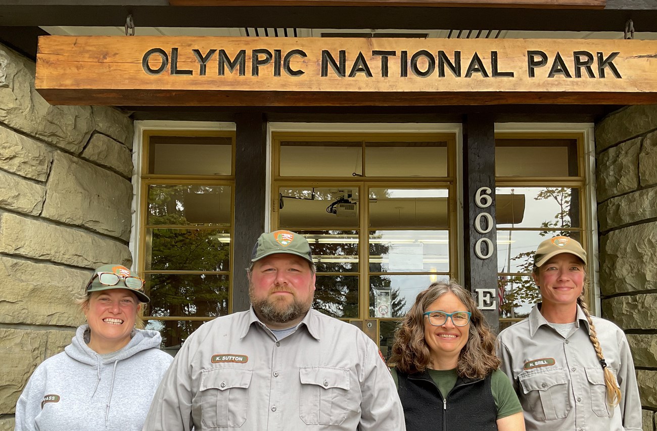 Four uniformed people stand smiling under the porch of a log building.