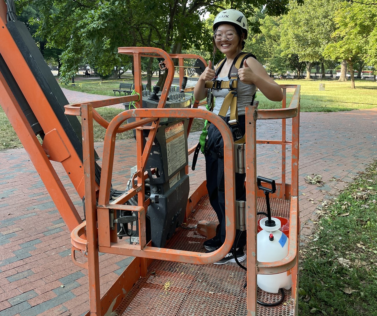 Jessica, an Asian American woman, stands on an orange aerial lift. Jessica is wearing a white hard hat. The aerial lift sits on a brick path.