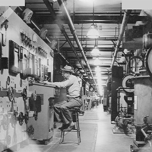 Operations Gallery A man sits on a tall stool along the left-hand side of a long hallway, adjusting a dial with his left hand. The walls are covered with dials, levers, and meters.