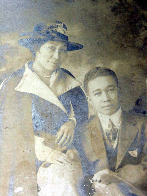 Black and white photo. A Filipino woman and man seated next to each other, looking into the camera. She is on the left, wearing a hat, dark top with a wide white collar. He is wearing a grey suit, collared shirt, tie, jacket, and a pocket square.
