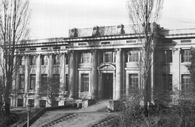 Black and white photo of a large campus building with columns and three floors of windows. The entrance is flanked by trees and approached by a large set of steps.