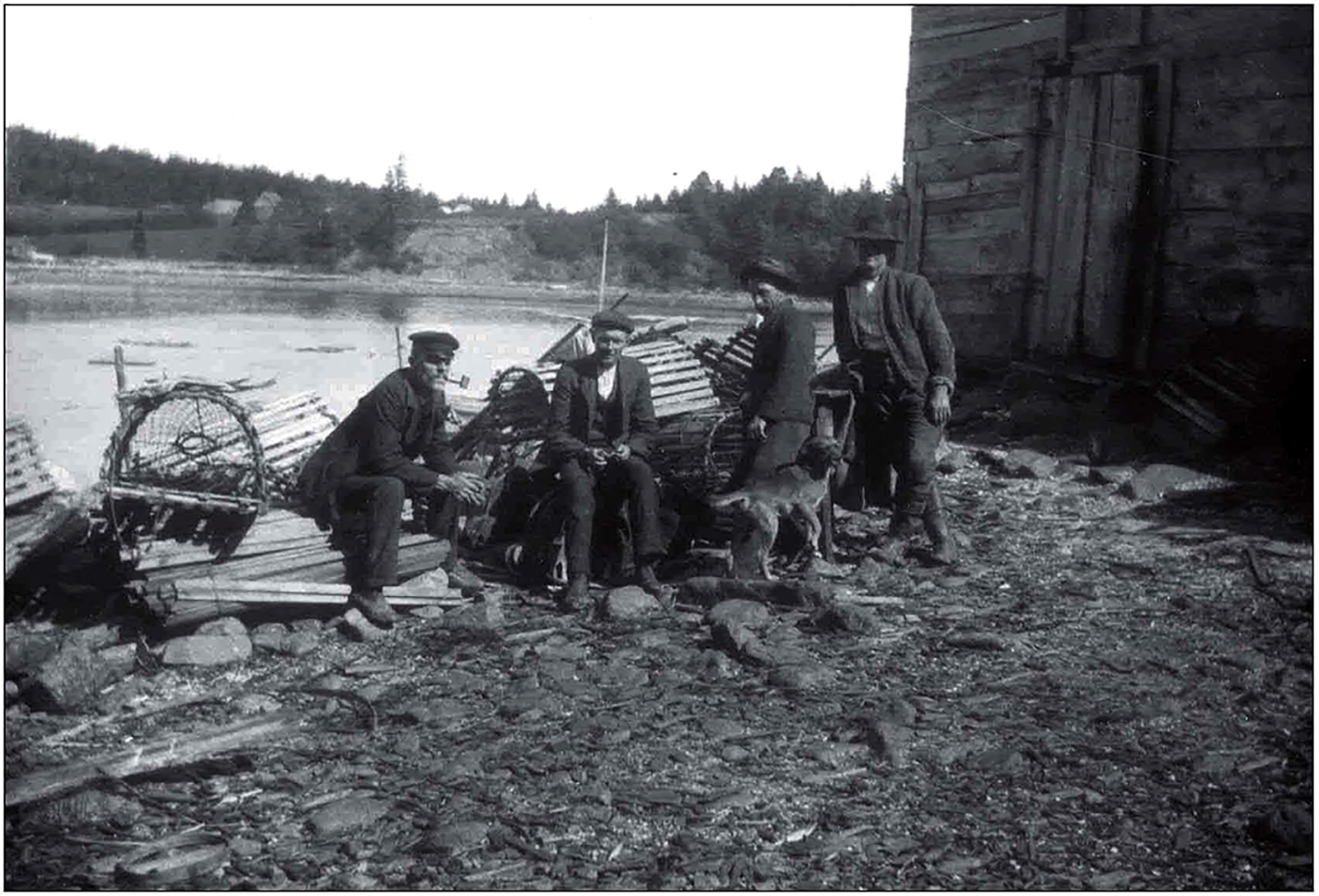 An old black and white photo of four men and a dog sitting or leaning against fishing gear in front of a waterway.