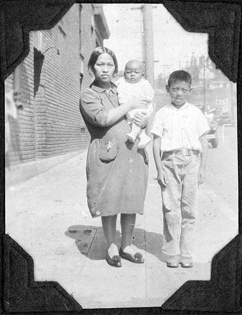 Home and Homelands Exhibition. Credit: Personal collection of Ou Shee and Art Wing Eng’s family photo album Black and white photo of a woman of Chinese origin in a dress. She is holding a baby. Next to her is a boy of Chinese descent in a white shirt and pants. They are on a city street with a brick building in the background.