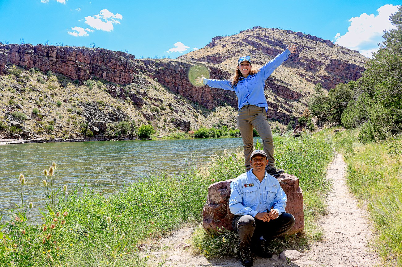 A man stoops below a rock while a woman stands on it with her hands outstretched above her. They are both smiling.