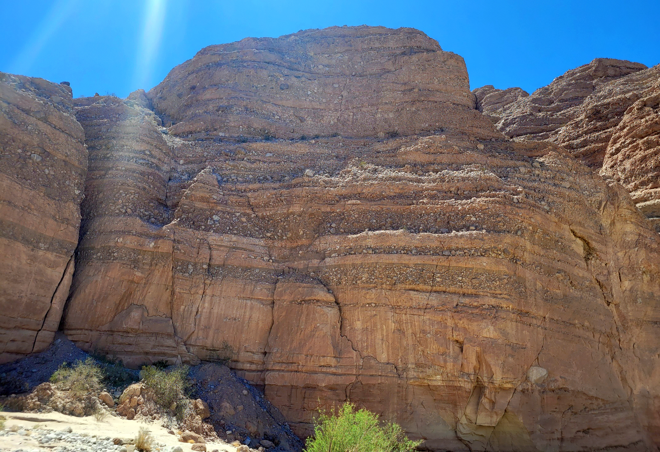 An large rock formation with multiple alternating layers of fine-grained rock and coarse, mixed rock with large cobbles, blue sky, sparse green vegetation