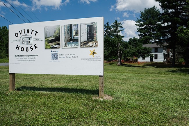 Photograph of a white large sign stands in front of the Oviatt House. The sign reads: Oviatt House Richfield Ohio Richfield Historical Preserve.