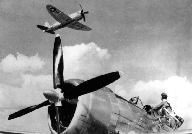 P-47 Thunderbolt fighter planes, one aloft, and the other on the ground with the pilot ready to climb into the cockpit. Both planes are light in color with a single front propeller.