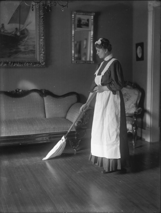 A maid in a black and white service uniform stands to the right, holding a broom.  She is sweeping a wood floor in front of a couch and chair and artwork on the walls.