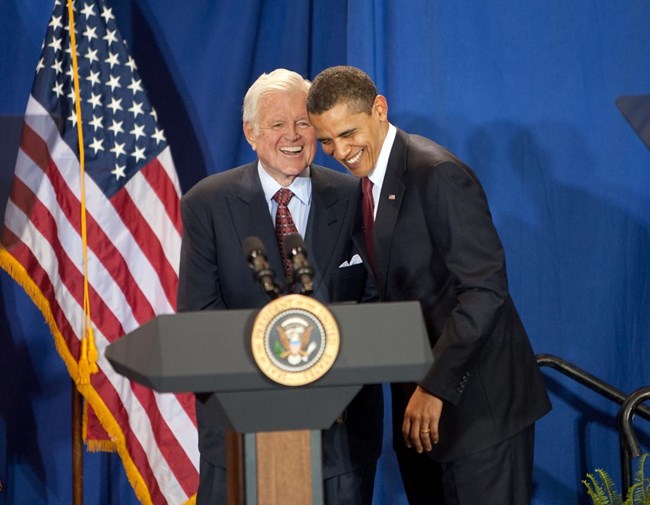 A color photo of Ted Kennedy behind a podium.  President Obama leans in from the right smiling.