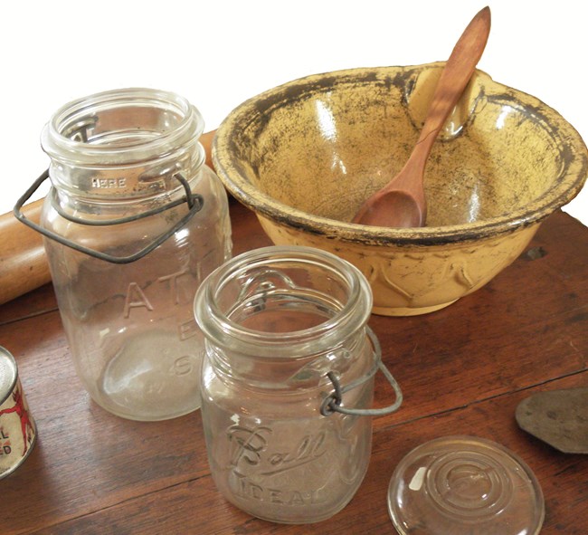 Two glass canning jars and a yellow bowl with a wooden spoon sit at the center of a wooden table.  On the left, a can of Underwood Deviled Ham is just visible as is a wooden rolling pin at the top of the photograph.