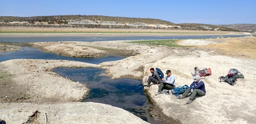 Three people sitting on bare, open ground next to a pool of water (spring emergence location) next to a narrow section of a lake in a very barren landscape.
