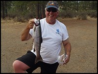 A man in a white t-shirt, shorts, and ball cap holds up a fish he caught.