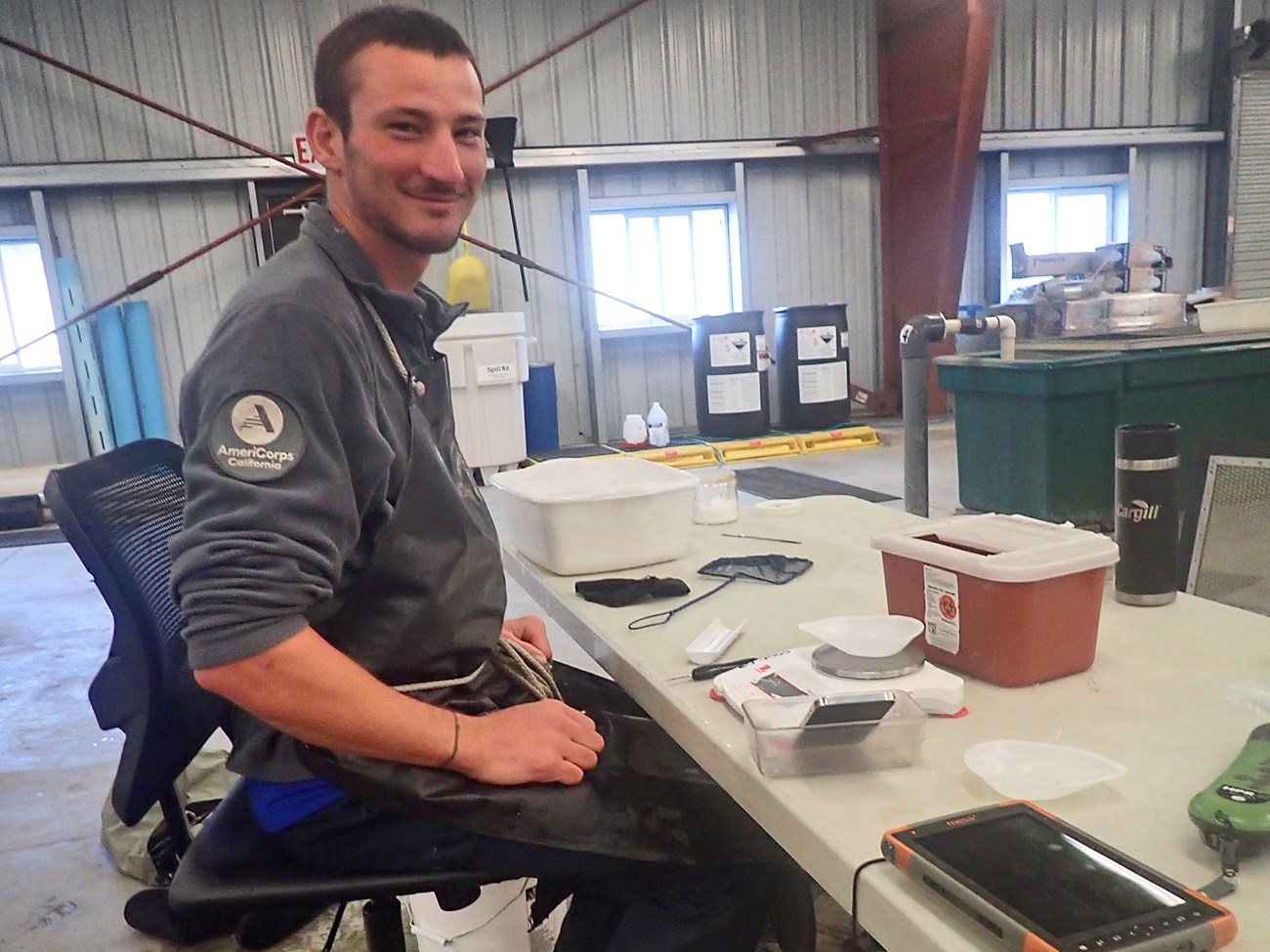 Val Kostelnik Kostelnik in a gray AmeriCoprps fleece and a black rubber apron, sitting at a table in a hatchery with equipment for netting, weighing, and tagging small fish.