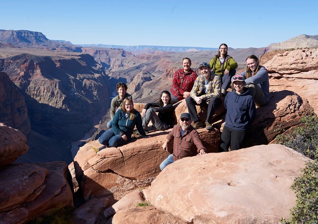 Nine people perch on large red-brown sandstone rocks overlooking the depths of Grand Canyon.