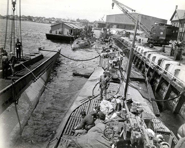 Four long, narrow submarines at a large dock, with people on and around them.