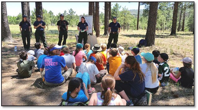 A group of children sit on the ground in a forest clearing. 5 wildland firefighters standing in front of the children