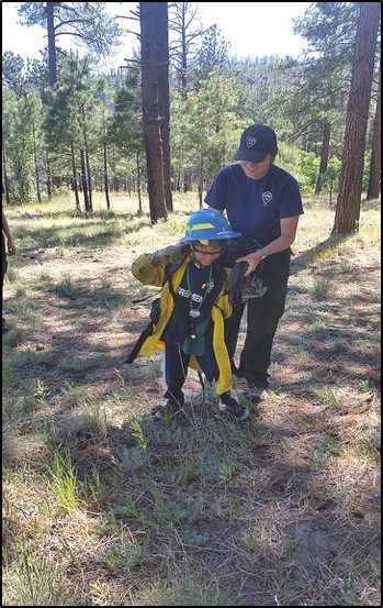 A woman assists a small child dressed in wildland fire gear, walking through a forest