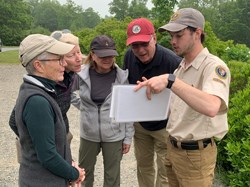 Ranger in a ball cap holds laminated images for a group of visitors gathered close around him. Ranger in a ball cap holds laminated images for a group of visitors gathered close around him.