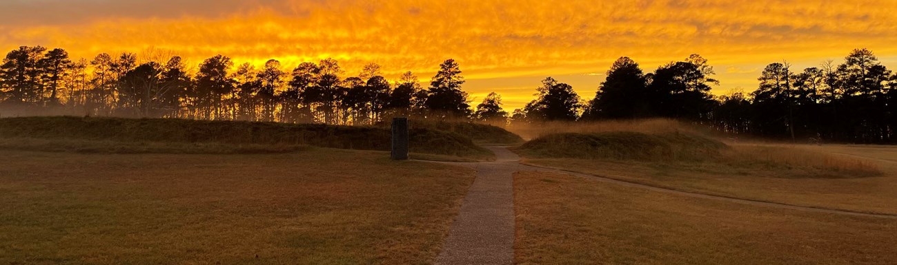Bright yellow-orange sunset silhouettes a line of trees on the horizon with the earthworks of Fort Stedman in the foreground. Bright yellow-orange sunset silhouettes a line of trees on the horizon with the earthworks of Fort Stedman in the foreground.