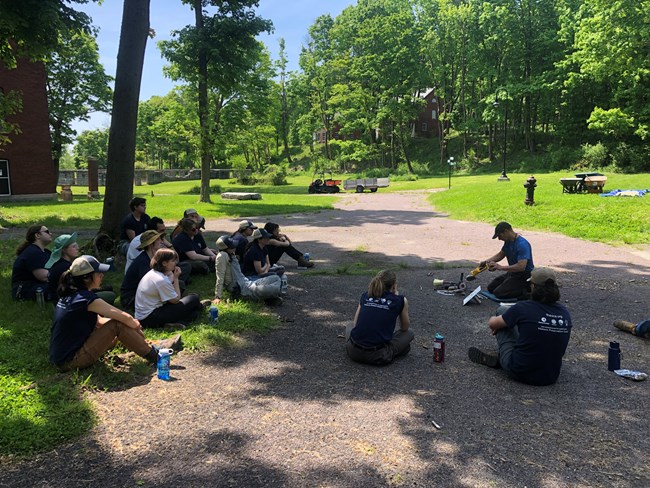 A group of corps members sitting on the ground, looking at a man sitting on the ground to the right. He is talking while looking down at an angle grinder in his hands, and there are several other tools on the ground in front of him.