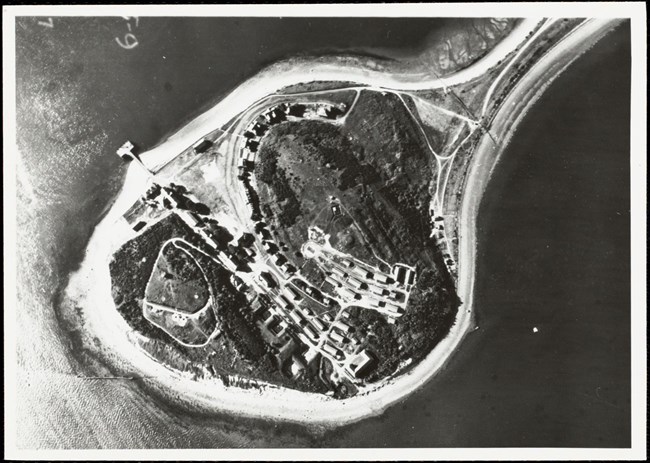 A black and white aerial photo of part of an island. The visible part of the island is circular and lined with a sandy beach, and is connected off-image to the rest of the island by a thin strip of land going into the top right corner of the image.