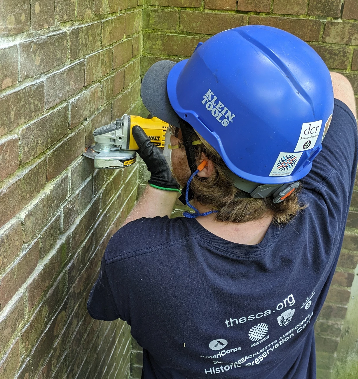 PIimg6_grinder A crew member wearing a hard hat, N95 mask, safety glasses, and earplugs holds an angle grinder up to an old brick wall, removing some of the mortar.