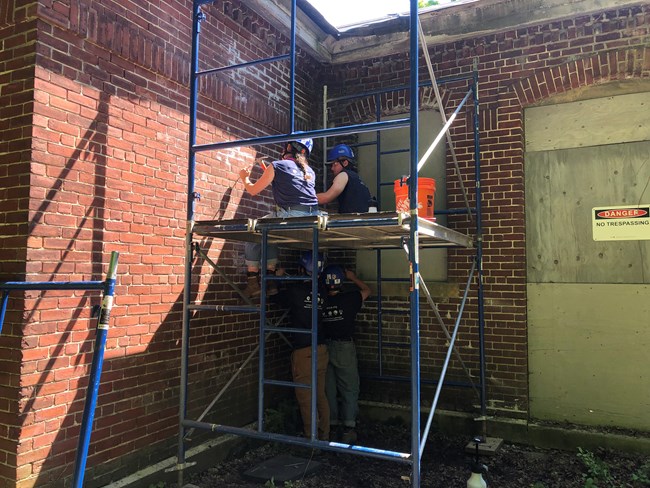 Scaffolding set up at the corner of an old brick building. There are four crew members wearing hard hats and holding hammers and chisels, two of them on the ground level and two of them sitting on the scaffolding platform above.