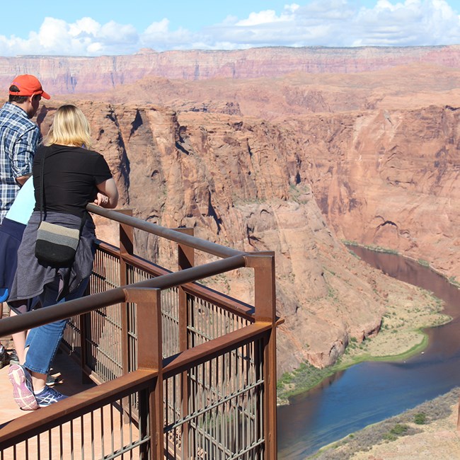 A few people lean against a overlook railing at the top of a canyon