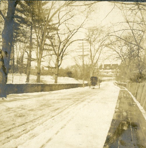 Historic image of a icy road with a horse drawn carriage in the distance with a large white building behind the carriage.