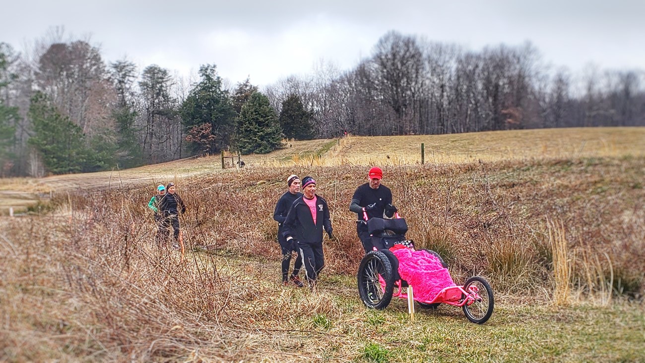 Event runners in a grass field including one pushing a wheel chair