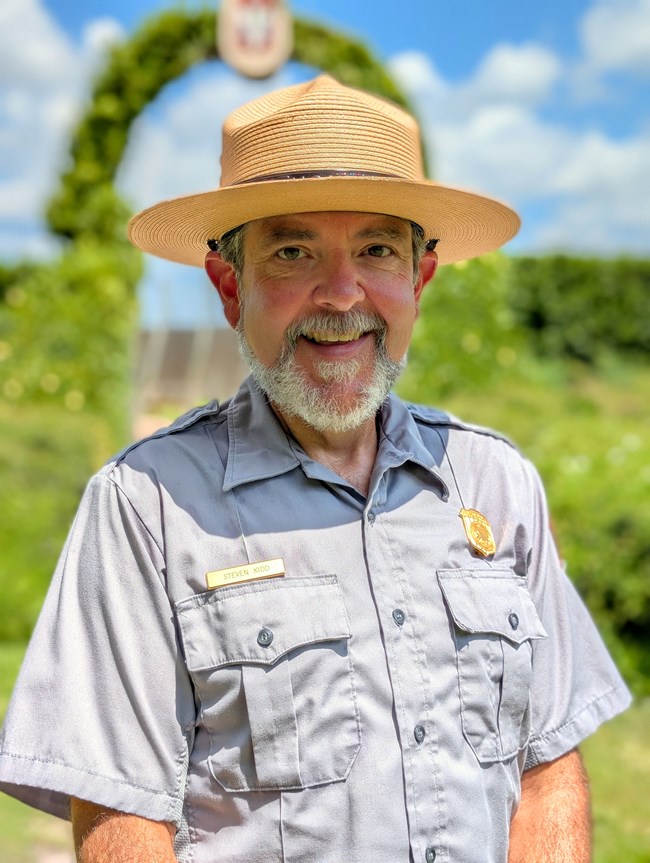 A tan-uniformed person with a wide-brimmed straw hat, short gray hair, mustache and trimmed beard with a big smile.
