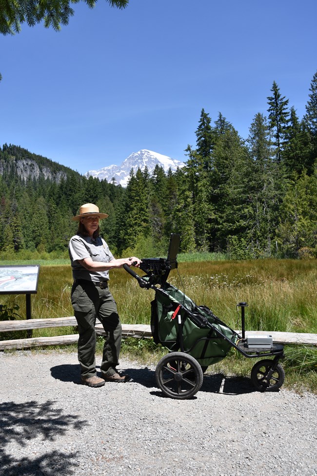 A uniformed person in a wide-brimmed straw hat holding the handlebars of a three-wheeled type of bike, in front of a landscape of large green trees and a distant snow-capped mountain.