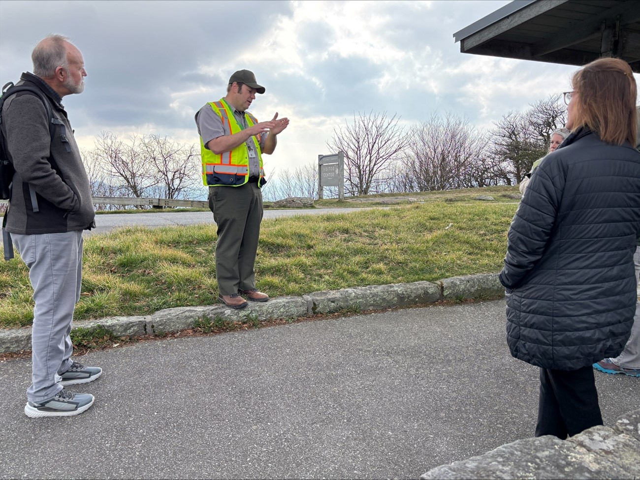 A person in a green ball cap, gray shirt, green pants, and orange safety vest stands near an outbuilding, talking to a few people.
