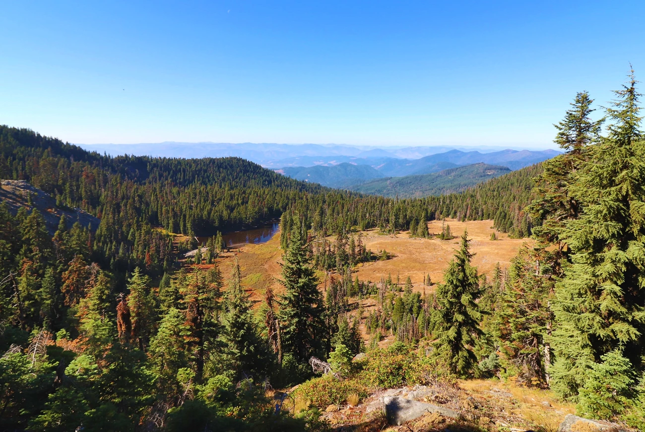 River Styx headwaters Looking down a mountain at a beautiful vista including evergreen trees, a golden meadow, a pond, and a range of lower mountains in the distance.
