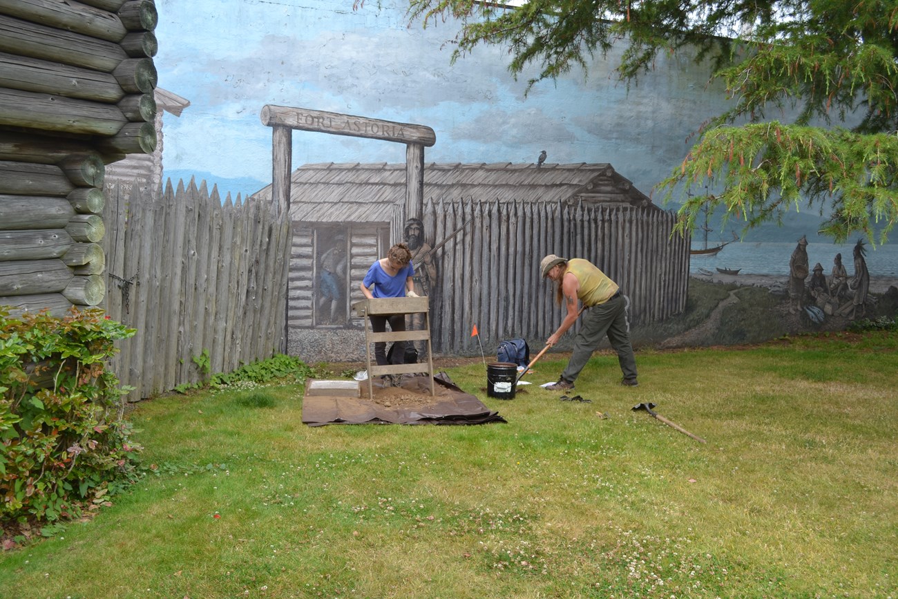 Two archeologists digging and sifting through soil by an outdoor mural of what Fort Astoria once looked like.