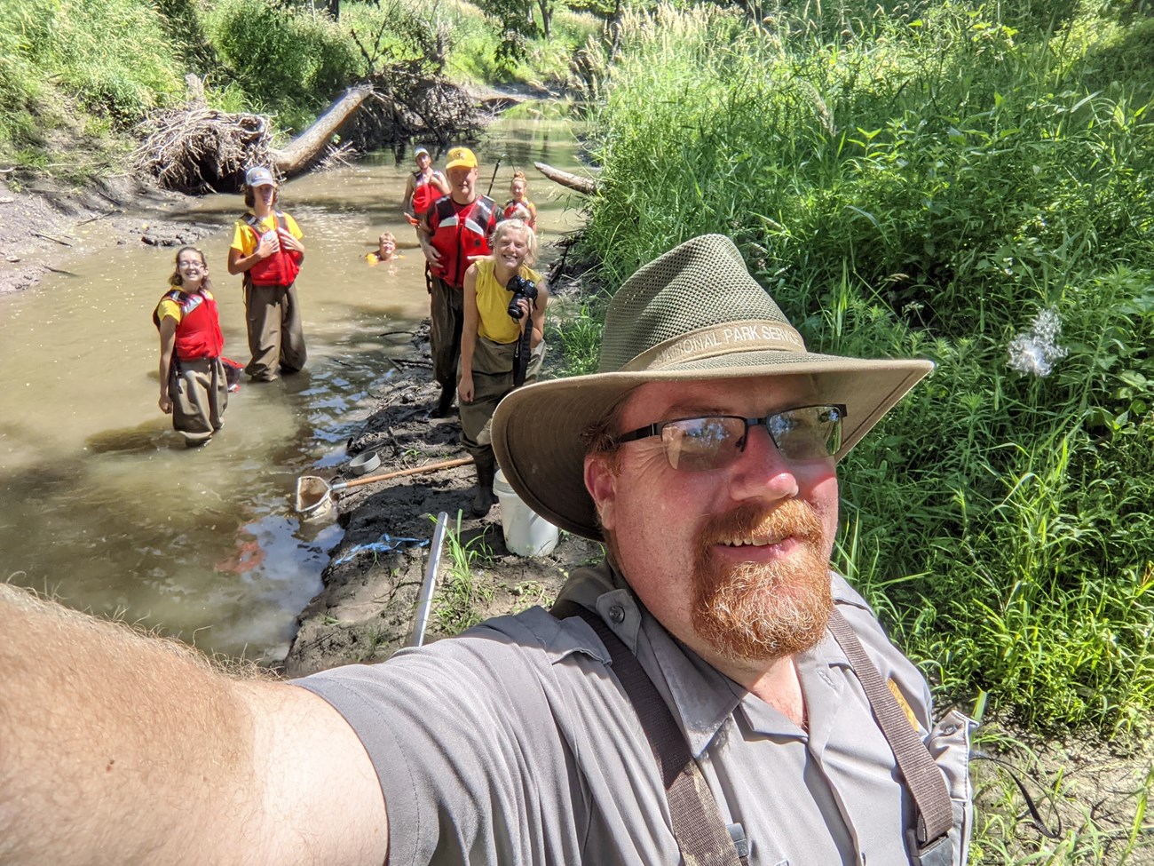Jesse Bolli with 8 Youth Conservation Corp members in Cub Creek. A D-net and a Secchi tube are on the muddy bank. Green vegetation grows up the sides.