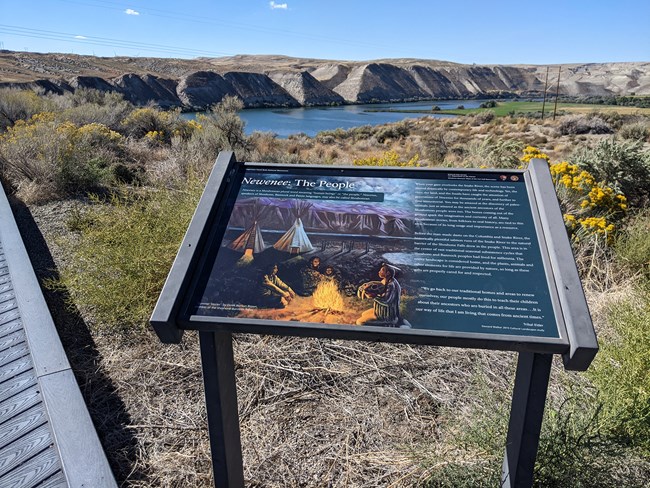 A wayside exhibit panel sits alongside a wooden boardwalk, with the Snake River and Fossil Beds in the background. The panel features Derek No-Sun Brown's painting, Summer Stories, and text.
