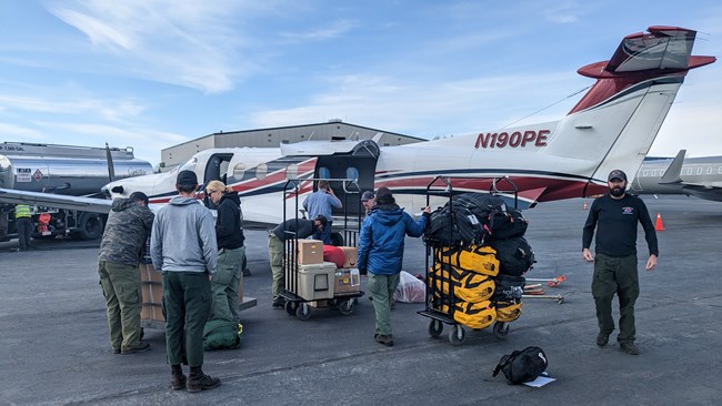 Several people stand in front of a small plane and wildland firefighting gear