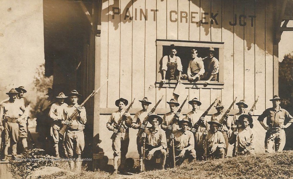 Striking miners during the Paint Creek Strike in 1912. A row of men in white shirts and hats stand near a wooden shack holding rifles.