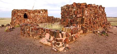 Two piles of rocks in reds and whites similar but larger than brick, arranged loosely into two dwellings, one with a window. A short perimeter of the same rocks appearing like a fence around the dwellings. Tall green grass grows inside he fence.