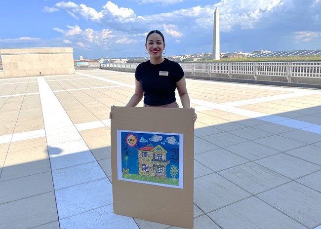 person holding a picture of a painting of a colorful old 1920s Mexican style house. The Washington Monument can be seen in the distance.