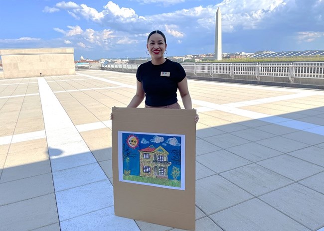 Paola Vazquez person holding a picture of a painting of a colorful old 1920s Mexican style house. The Washington Monument can be seen in the distance.