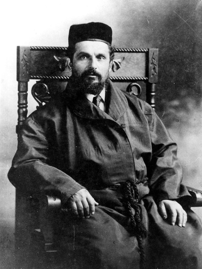 Rabbi Benjamin Papermaster Black-and-white photograph of a bearded man seated in a wood chair with decorative carvings. He is wearing a Bukharian kippah and a dark robe with a tassel belt, worn over a shirt and tie.