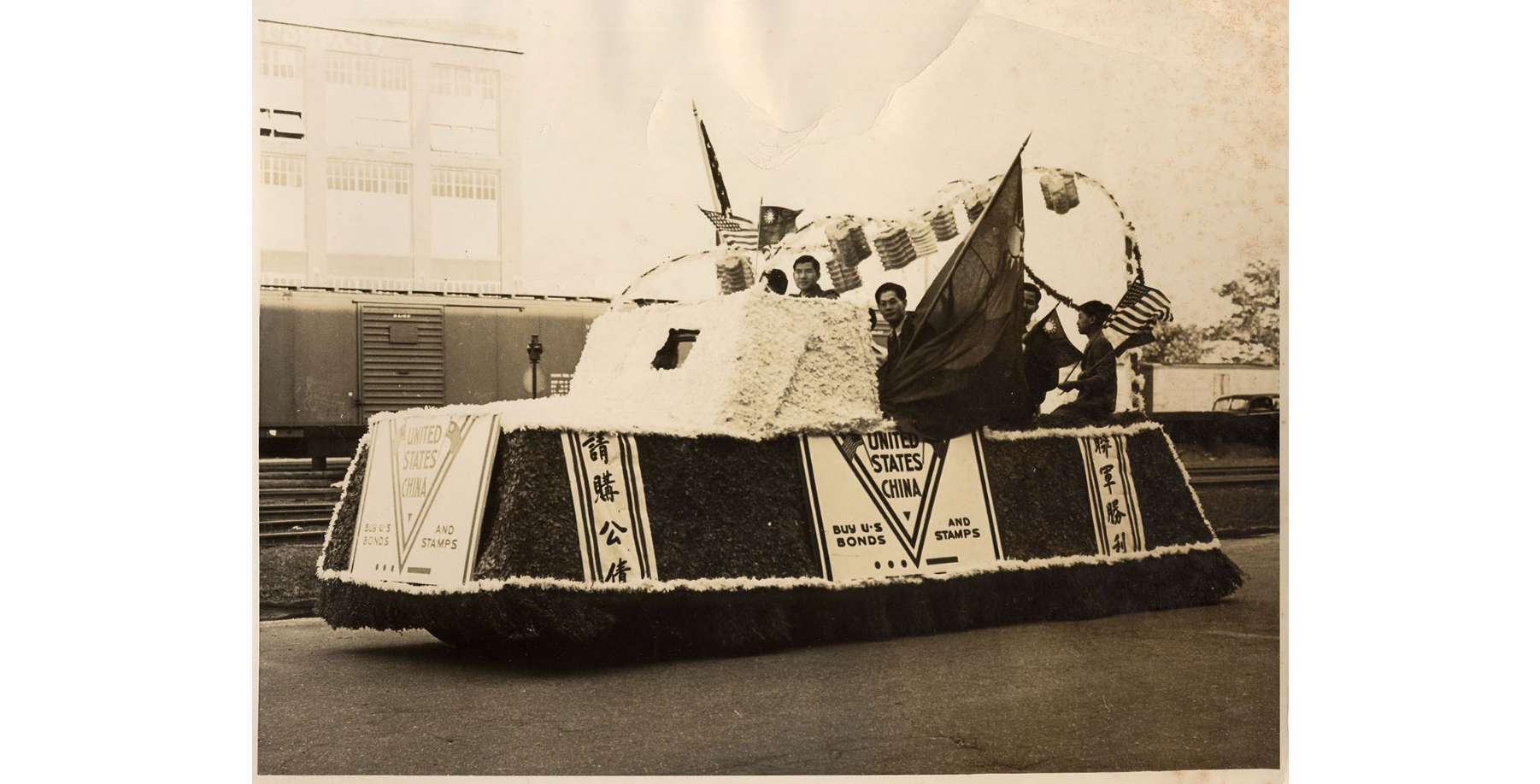 World War II Parade Float Black and white photo of a paper machier covered car. Flags are hanging from the top. A few men of Asian descent are visible sitting in the bed of the car.
