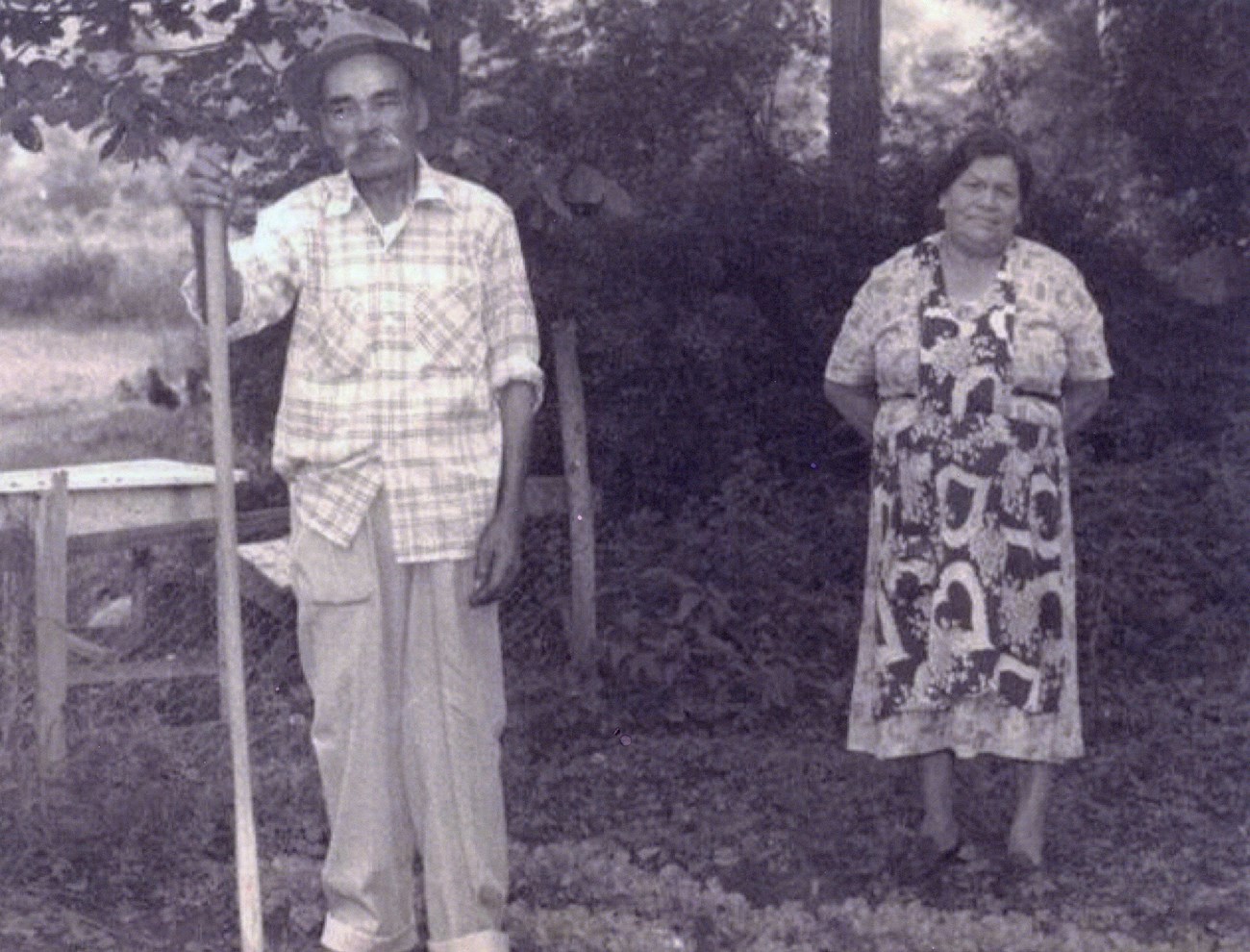 Black and white photo of a couple standing in a yard next to a chicken pen and trees. He has a bushy mustache and is dressed in a checked shirt, loose pants, and a hat. He leans on a staff. She wears an apron with large hearts over a patterned dress.