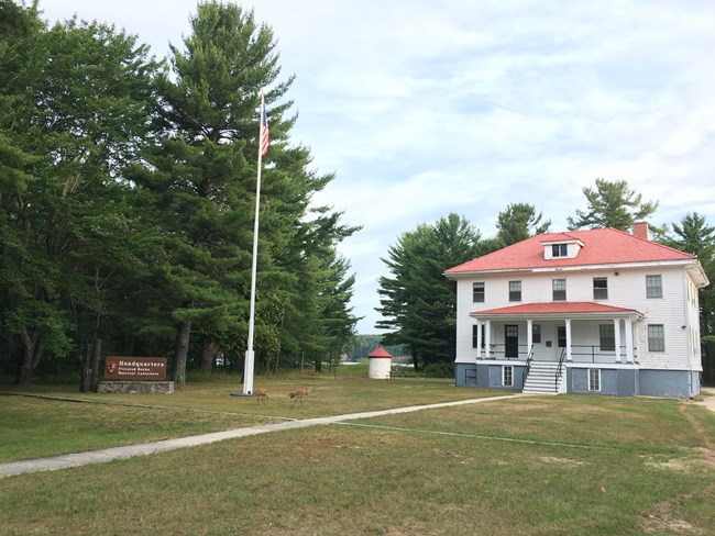 Park Headquarters at Sand Point in Munising on a summer day. Park sign is in front next to the flag pole. A pair of sandhill cranes are on the lawn.