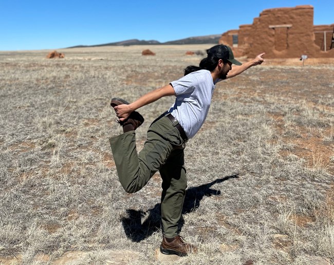 Park ranger doing Lord of the Dance yoga pose, standing on their left leg and leaning their body to the left while extending their left arm to the left. Their right leg is lifted upward and their right foot is held by the fully extended right arm.