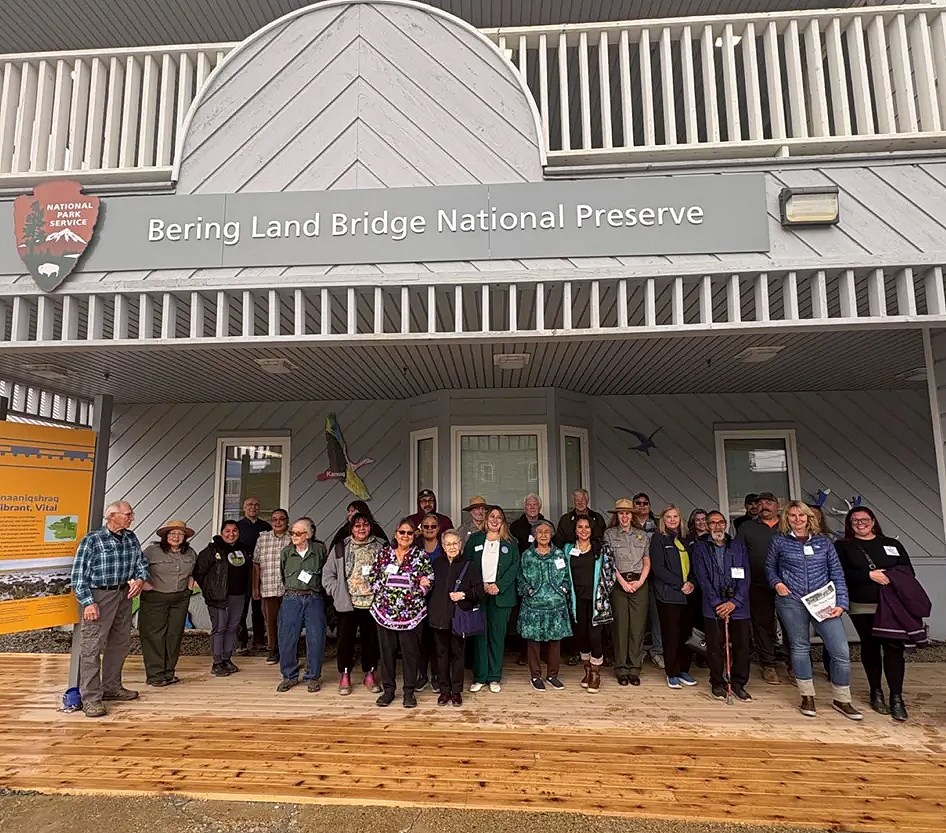 A group of over 15 people stand together in a building's wooden porch under a white portico.