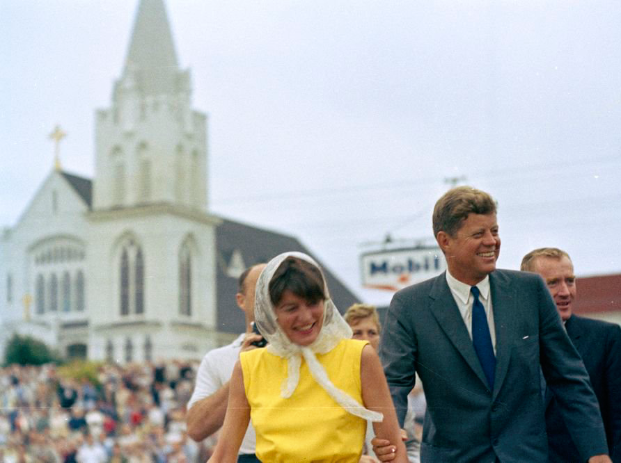 A photo of Patricia Kennedy Lawford, in a bright yellow sleeveless dress and lace veil, her arm held by President Kennedy, in a grey suit and blue tie. They are both smiling. A large crowd is gathered around a white church in the background.